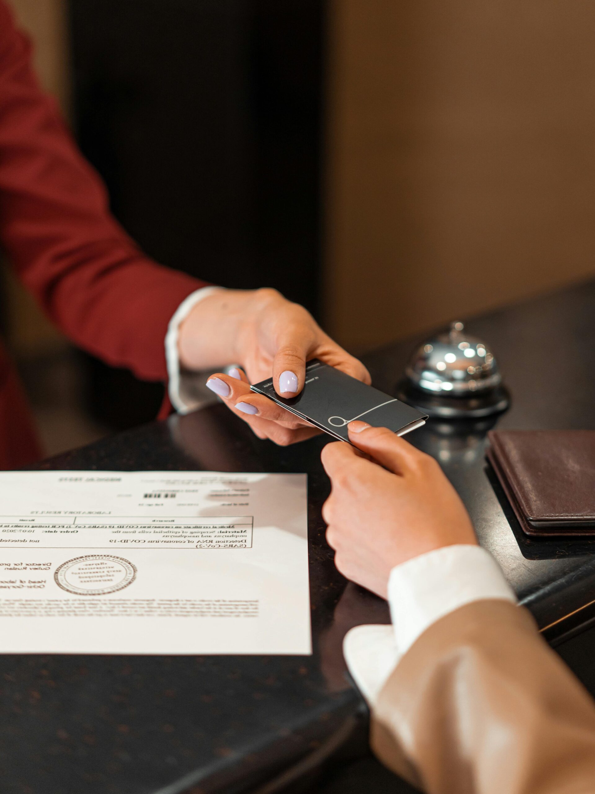 A hotel receptionist hands a key card to a guest over the counter, with documents visible.